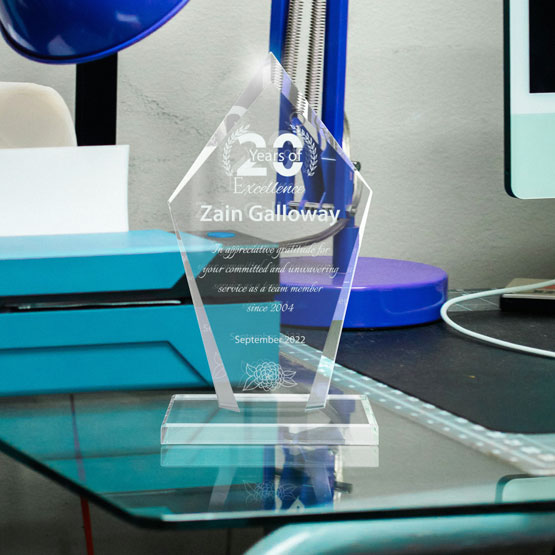 A clear glass retirement award, engraved with personalized white text and a floral design, displayed on glass desk in an office setting.