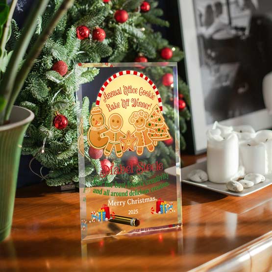 A clear acrylic holiday keepsake award standing on a wooden table, featuring festive gingerbread illustrations and Christmas-themed text. The background shows a decorated Christmas tree, candles, and framed wall art in a home setting.