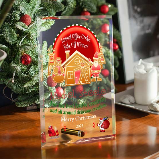 A clear acrylic holiday keepsake award standing on a wooden table, featuring festive gingerbread illustrations and Christmas-themed text. The background shows a decorated Christmas tree, candles, and framed wall art in a home setting.