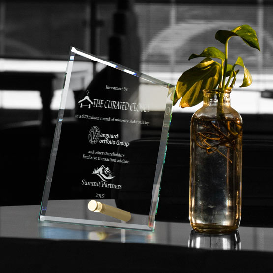 A clear glass award engraved with text and logos, standing on a reflective dark surface beside a small glass vase with a green plant in a real indoor setting.