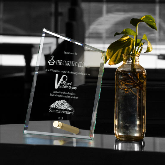 A clear glass award engraved with text and logos, standing on a reflective dark surface beside a small glass vase with a green plant in a real indoor setting.