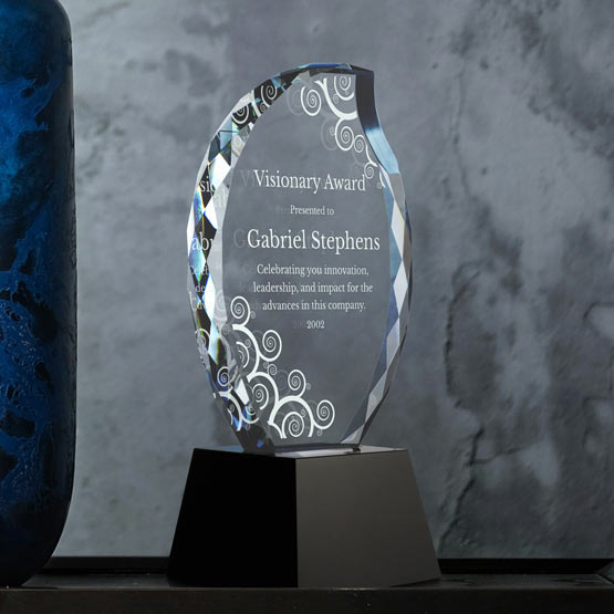 A clear glass trophy award on a black pedestal base, engraved with the text ‘Visionary Award presented to Gabriel Stephens’ and decorative floral motifs, set against a gray textured wall with a blue vase partially  visible on the left.