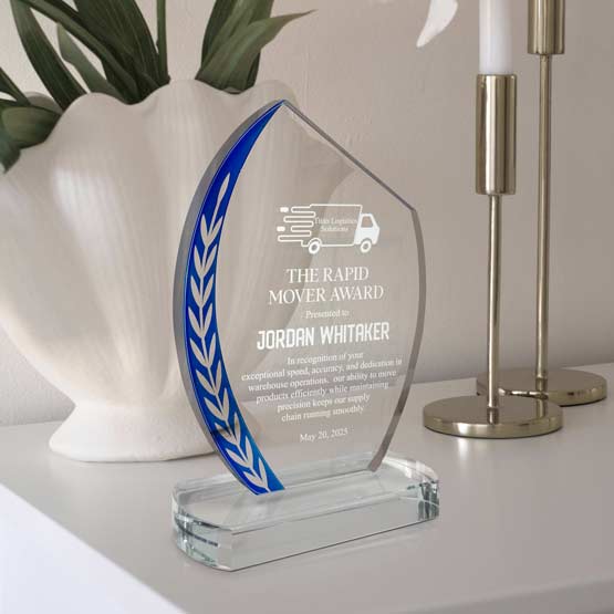 Custom Slanted Oval Glass Award For Moving Employee With Blue Edge And Glass Base On White Shelf With Lamps And Potted Plant