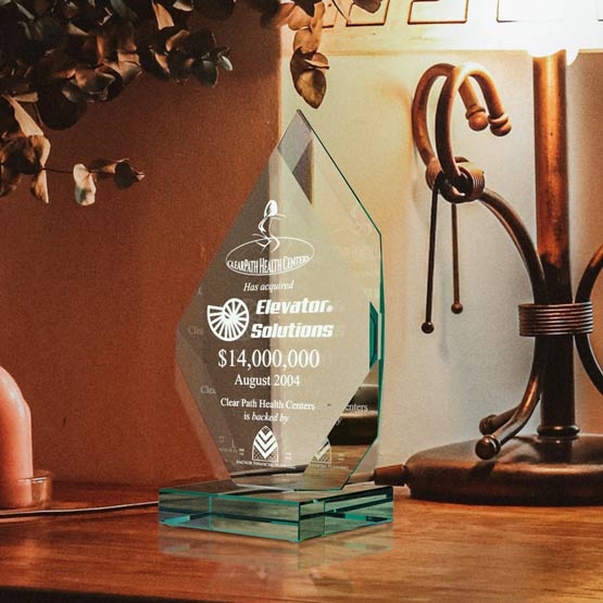 A jade tinted glass award with engraved text and graphics, displayed on a wooden table next to a lit lamp and decorative branches.
