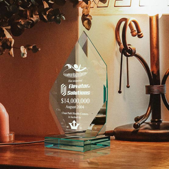 A jade tinted glass award with engraved text and graphics, displayed on a wooden table next to a lit lamp and decorative branches.