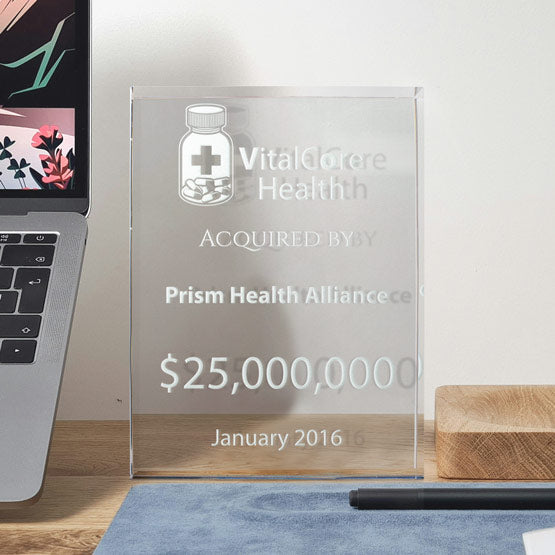 A rectangular clear glass award with engraved frosted text saying ‘VitalCore Health Acquired by Prism Health Alliance’ with a large dollar amount. Displayed on a wooden text with a laptop next to the award and blue mat at the bottom.