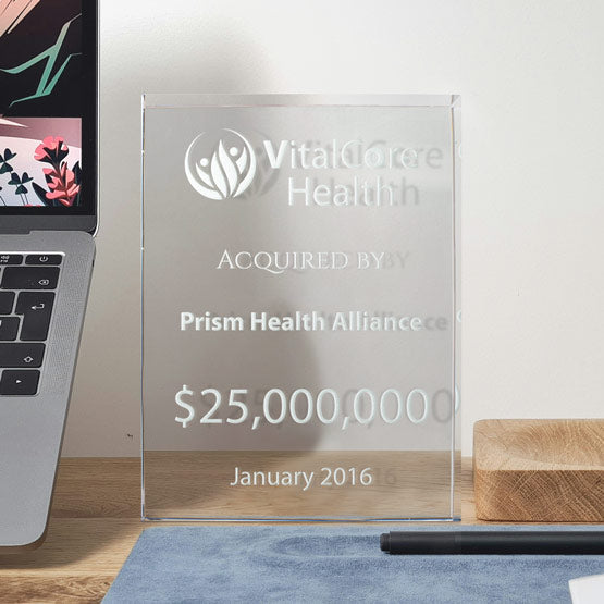 A rectangular clear glass award with engraved frosted text saying ‘VitalCore Health Acquired by Prism Health Alliance’ with a large dollar amount. Displayed on a wooden text with a laptop next to the award and blue mat at the bottom.
