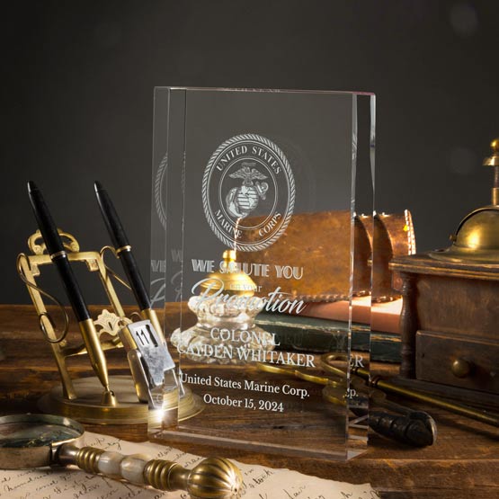 A clear glass personalized military award plaque engraved with the United States Marine Corps emblem and text, displayed on a wooden desk with brass stationery and a vintage brass clock in the background.