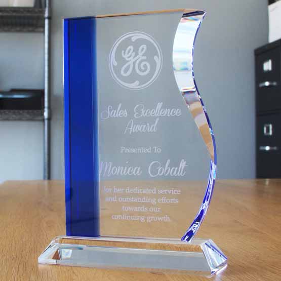 Custom Wave Shaped Glass Award For Sales Excellence With Blue Edge And Clear Glass Base On Wood Office Desk Near Shelves