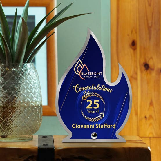 A custom flame-shaped stainless steel award celebrating 25 years, featuring a printed deep blue center with gold and white lettering, displayed on a wooden surface next to a potted plant.