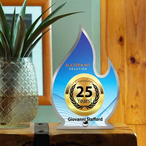 A custom flame-shaped stainless steel award celebrating 25 years, featuring a printed deep blue center with gold and white lettering, displayed on a wooden surface next to a potted plant.