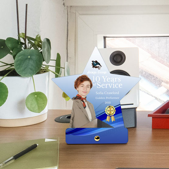 A custom star-shaped stainless steel award displayed on a wooden desk in an office setting. The award features a photo of a woman, the text recognizes 10 years of service, with a gold and teal design with gold pin at the bottom for mounting.