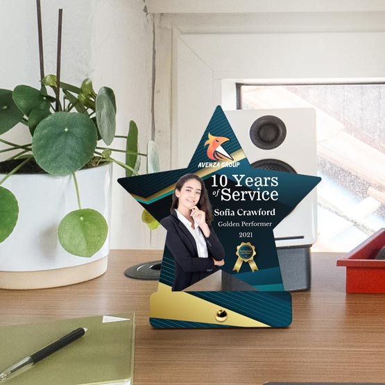 A custom star-shaped stainless steel award displayed on a wooden desk in an office setting. The award features a photo of a woman, the text recognizes 10 years of service, with a gold and teal design with gold pin at the bottom for mounting.