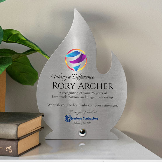 A flame-shaped stainless steel metal award with color printed retirement recognition text and multicolor logo, displayed on a tabletop with a silver pin next to stacked books and a green plant against a neutral wall.