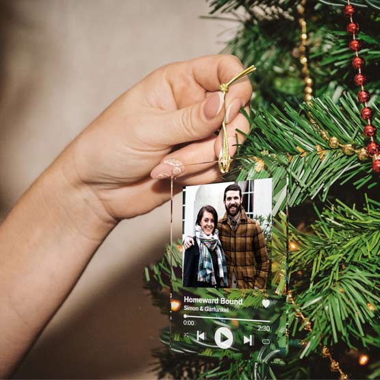 A hand hangs a clear acrylic rectanglar music-player style ornament from a green Christmas tree branch. The ornament displays a photo of a couple and the text ‘Homeward Bound Simon & Garfunkel’ with playback controls and a gold hanging loop.
