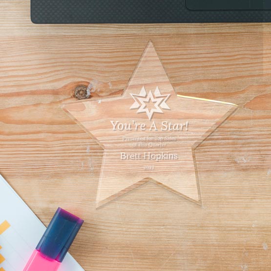 A clear acrylic star-shaped award engraved with ‘You’re A Star!’ and personalized text, displayed on a light wooden desk next to a laptop and highlighter.
