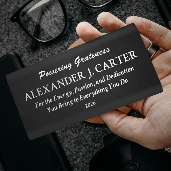 Hand Holding A Black Aluminum Power Bank Custom Engraved With Name And Appreciation Message Above Office Desk And Glasses