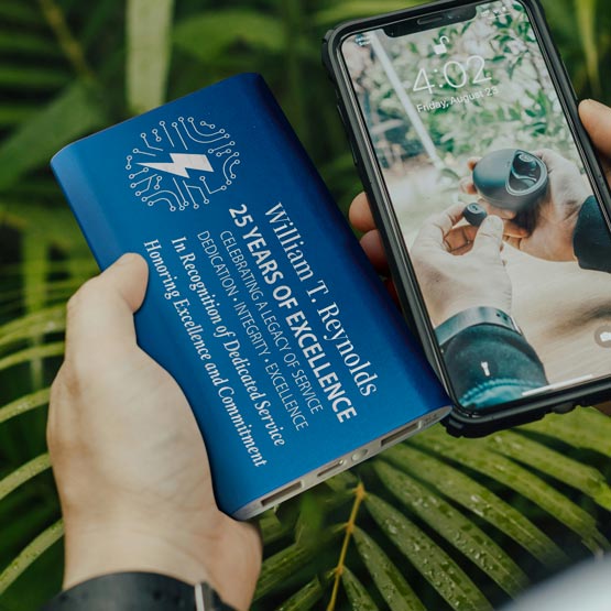 Hands Outdoors Holding A Blue Aluminum Power Bank Custom Engraved With Tech Logo And Years Of Service Award Message 