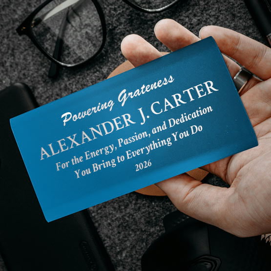 Hand Holding A Blue Aluminum Power Bank Custom Engraved With Name And Appreciation Message Above Office Desk And Glasses