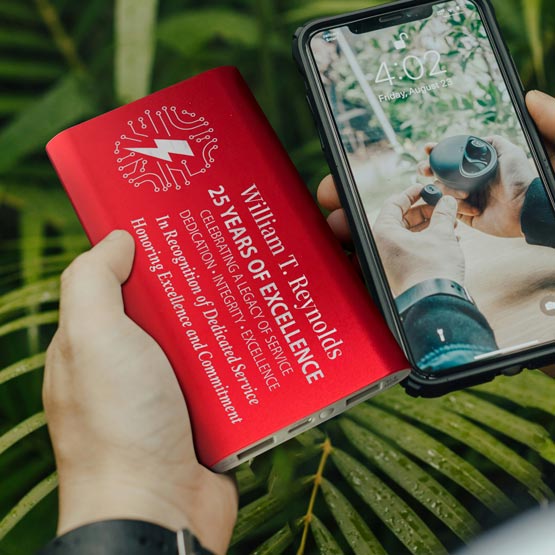 Hands Outdoors Holding A Red Aluminum Power Bank Custom Engraved With Tech Logo And Years Of Service Award Message