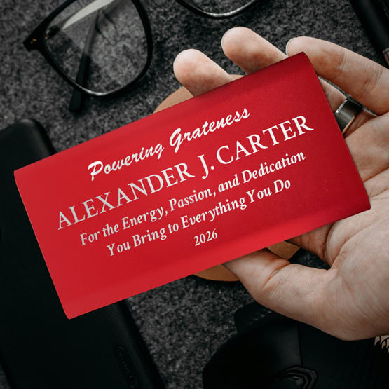 Hand Holding A Red Aluminum Power Bank Custom Engraved With Name And Appreciation Message Above Office Desk And Glasses