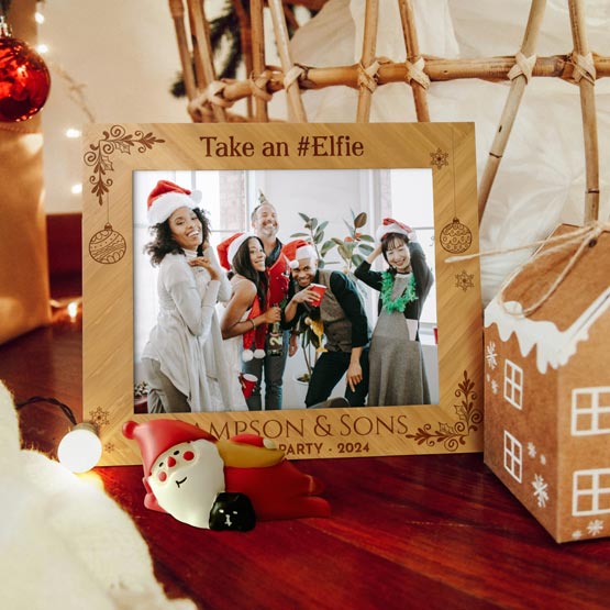 A personalized bamboo wooden Christmas party photo frame with the text ‘Take an #Elfie’ at the top and ‘Simpsons & Sons Party 2024’ at the bottom. Styled on a wooden table with holiday decorations including a gingerbread house, Santa figurine, and warm string lights.