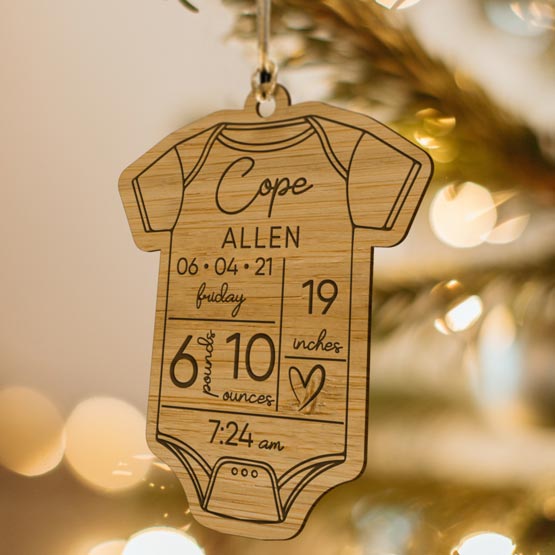 Custom Onesie Shaped Bamboo Ornament Engraved With Allen Cope And Baby Birth Details Hanging On Lit Christmas Tree