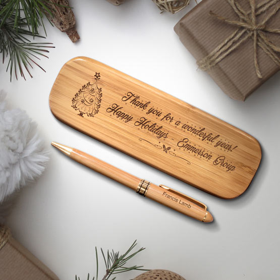 A bamboo pen case with engraved text on the front that reads ‘Thank you for a wonderful year! Happy Holidays Emmerson Group’. A matching pen sits right next to the case with a name engraved on the pen reading ‘Francis Lamb’. The set is on a white table surrounded by rustic looking Christmas themed props including a brown paper wrapped gifts and evergreen branches.