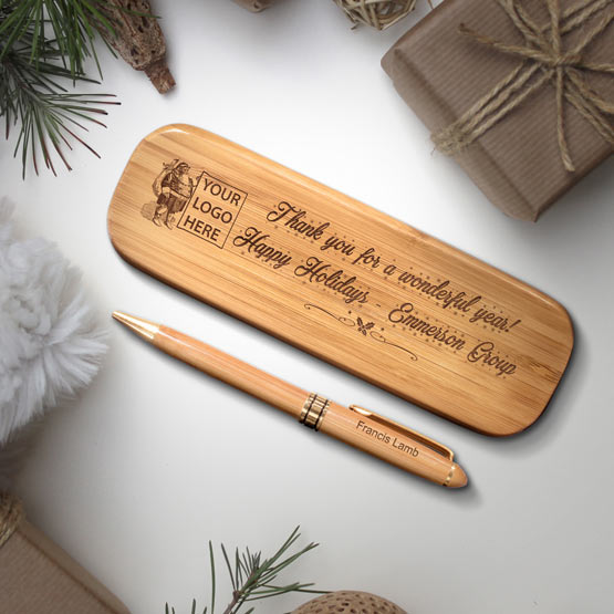 A bamboo pen case with engraved text on the front  with a holiday message. A matching pen sits right next to the case with a name engraved on the pen reading ‘Francis Lamb’. The set is on a white table surrounded by rustic looking Christmas themed props including a brown paper wrapped gifts and evergreen branches.