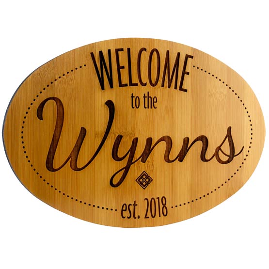 Custom Oval Shaped Bamboo Sign Laser Engraved With Welcome To The Wynns Text And Date Set Against White Background