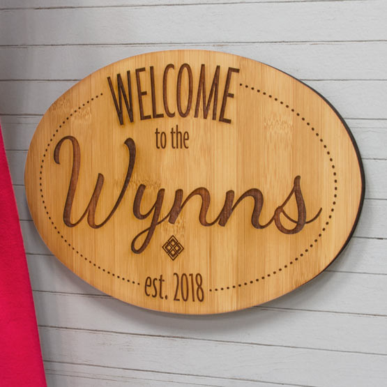Custom Oval Bamboo Sign Laser Engraved With Welcome To The Wynns Text And Date Hanging On House Siding