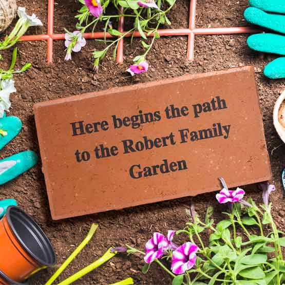 A red brick engraved with the text ‘Here begins the path to the Robert Family Garden’, styled on soil with gardening gloves, flowers, and pots.