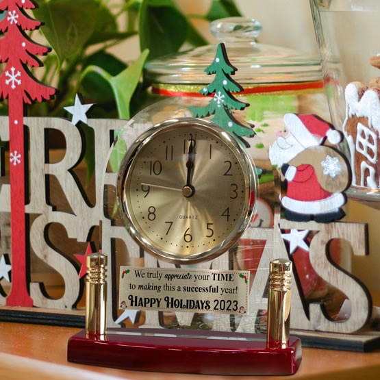 A arch crystal clock with a gold metal tag attached to the bottom and black numerals, set on a rosewood base and gold post. The clock is decorated with a small green Christmas tree topper and placed in front of a holiday-themed wooden sign and glass jar.