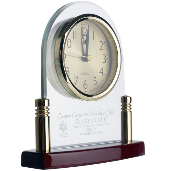 A glass clock with a clear arched glass body, gold-toned bezel and post, a cream dial with black numerals, and a glossy dark rosewood base. The glass has frosted text engraved on the glass, set on a white background.