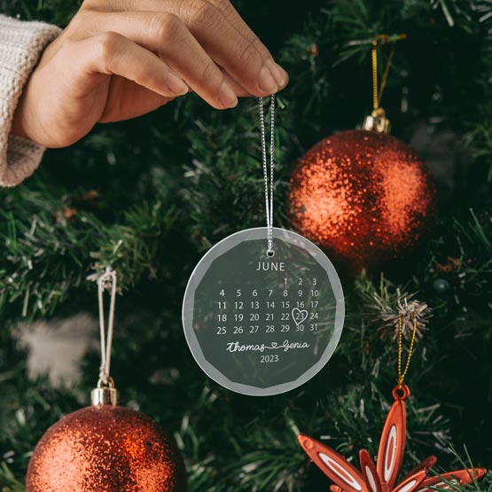 Hand Hanging A Custom Circle Glass Ornament Engraved With Calendar Art And Couple Names And Date Onto Christmas Tree