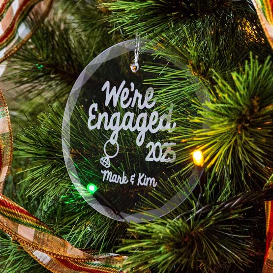 Custom Circle Glass Ornament Engraved With We’re Engaged Text And Date And Couple Names And Ring Art On A Christmas Tree