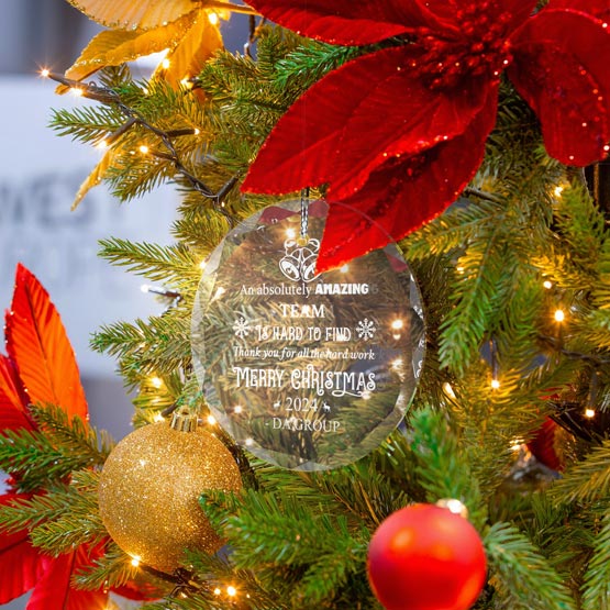A round glass ornament hanging from a silver string, engraved with festive text and snowflake details. Displayed on a decorated Christmas tree with green branches, red flowers, gold and red baubles, and warm string lights.