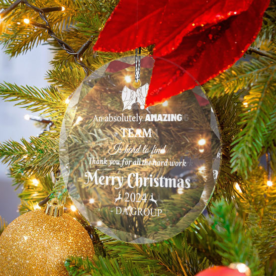 A circle glass ornament hanging from a decorated Christmas tree with string lights. Engraved text about a corporate team and their amazing work for the year.