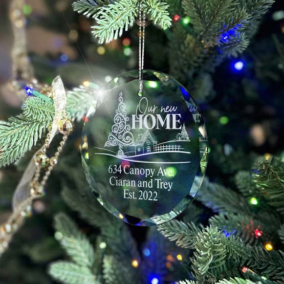 A clear glass circle ornament hanging from a silver string on a fully decorated Christmas tree with cool tone mostly blue string lights. The ornament is engraved with a graphic of decorative Christmas tree design in the foreground with a tiny house in the background, with text that reads ‘634 Canopy Ave. Ciaran and Trey Ets. 2022’.