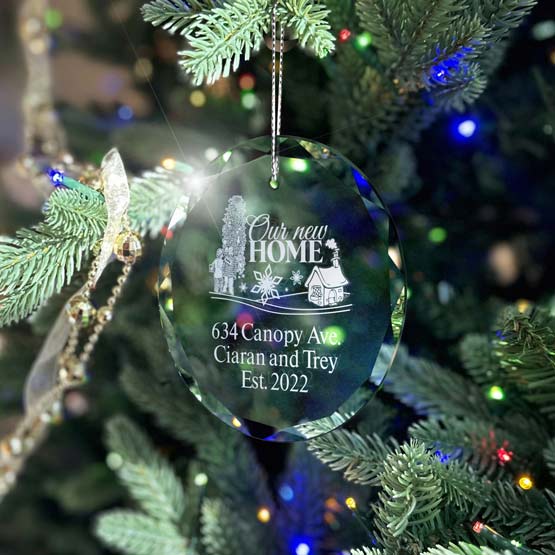 A clear glass circle ornament hanging from a silver string on a fully decorated Christmas tree with cool tone mostly blue string lights. The ornament is engraved with a graphic of decorative Christmas tree design in the foreground with a tiny house in the background, with text that reads ‘634 Canopy Ave. Ciaran and Trey Ets. 2022’.