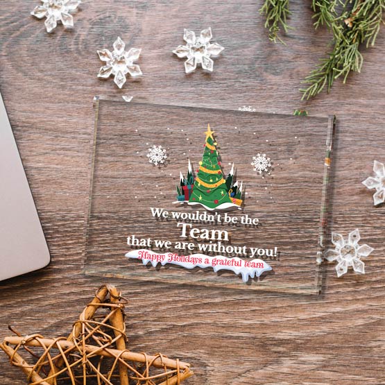 A clear glass paperweight displaying a colorful festive printed design featuring a Christmas tree graphic with text reading ‘We wouldn’t be the Team that we are without you!’. Laying on a wooden desk surrounded my decorative snowflakes, a wooden star and a laptop off to the left.