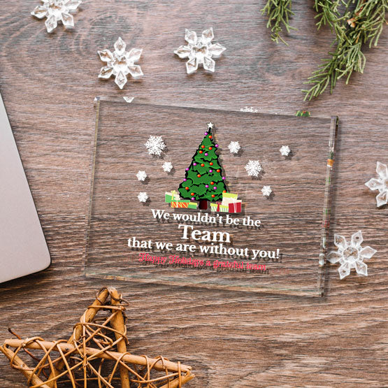 A clear glass paperweight displaying a colorful festive printed design featuring a Christmas tree graphic with text reading ‘We wouldn’t be the Team that we are without you!’. Laying on a wooden desk surrounded my decorative snowflakes, a wooden star and a laptop off to the left.