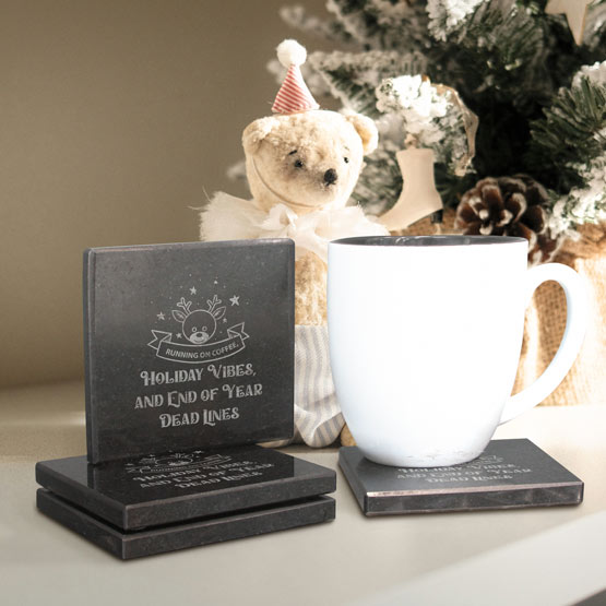 A set of granite engraved coasters, including one standing coaster that reads ‘HOLIDAY VIBES AND END OF YEAR DEAD LINES’ with a reindeer graphic, shown alongside a white mug. A teddy bear wearing a party hat and a decorated Christmas tree in the background.