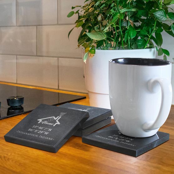 A white ceramic mug with a black interior sits on a granite stone square coaster on a wooden countertop. Two additional matching coasters are stacked behind it, and a potted green plant and tiled kitchen wall are visible in the background.