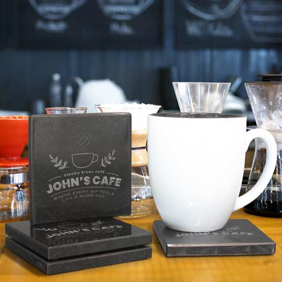 Set Of Four Custom Square Granite Coasters Engraved With John’s Cafe Text And Logo On Countertop Beside Coffee Mug