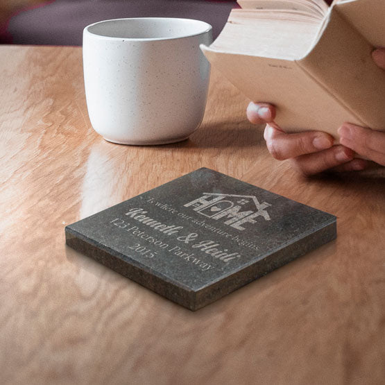 A personalized granite engraved stone coaster on a wooden table next to a white ceramic and a person holding a open book.