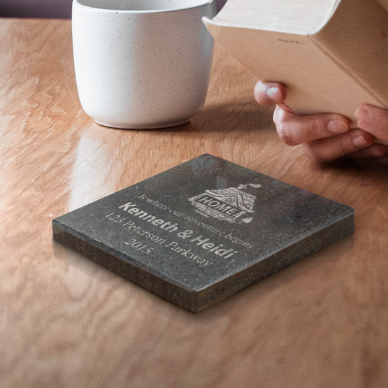 A personalized granite engraved stone coaster on a wooden table next to a white ceramic and a person holding a open book.