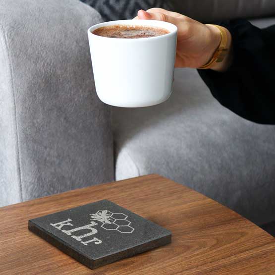 Woman Hand Placing Coffee Mug Onto Custom Square Granite Coaster With Light Flecks And Engraved With Logo Featuring Bee Art 