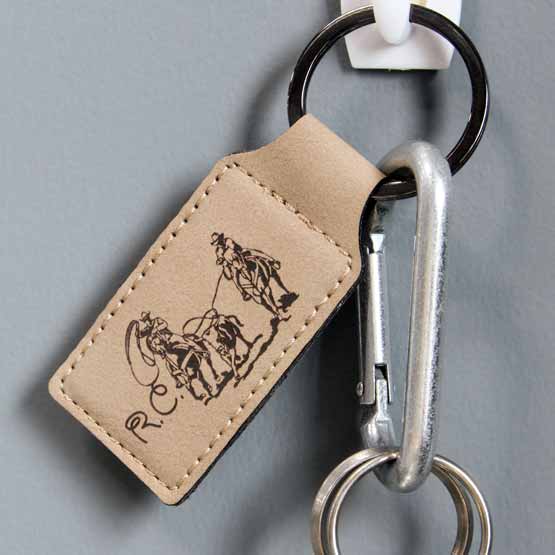 Light Brown Faux Leather Rectangle Keychain Engraved With Initials And Cowboys Lassoing Cow Artwork Hanging On A Blue Wall 