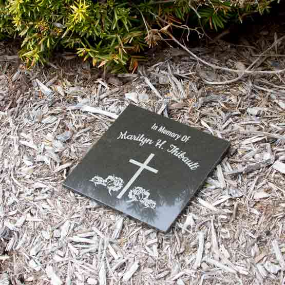 Square Black Marble Tile Engraved With Cross And Flower Art And In Memory Of Marilyn Thibault Text Laying Outside On Mulch
